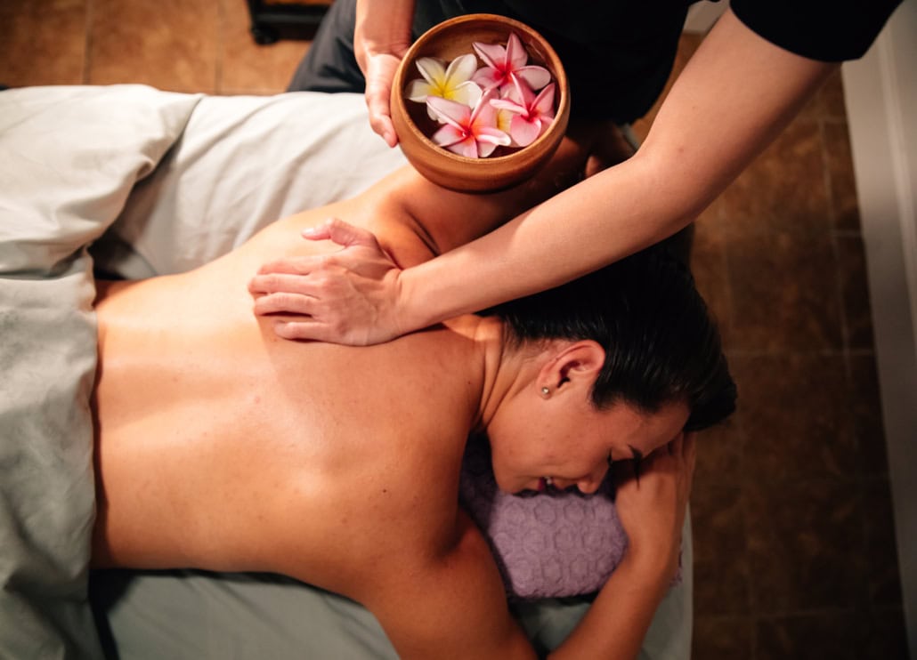 A woman, face down, getting a massage. The masseuse is holding a bowl of plumeria flowers.