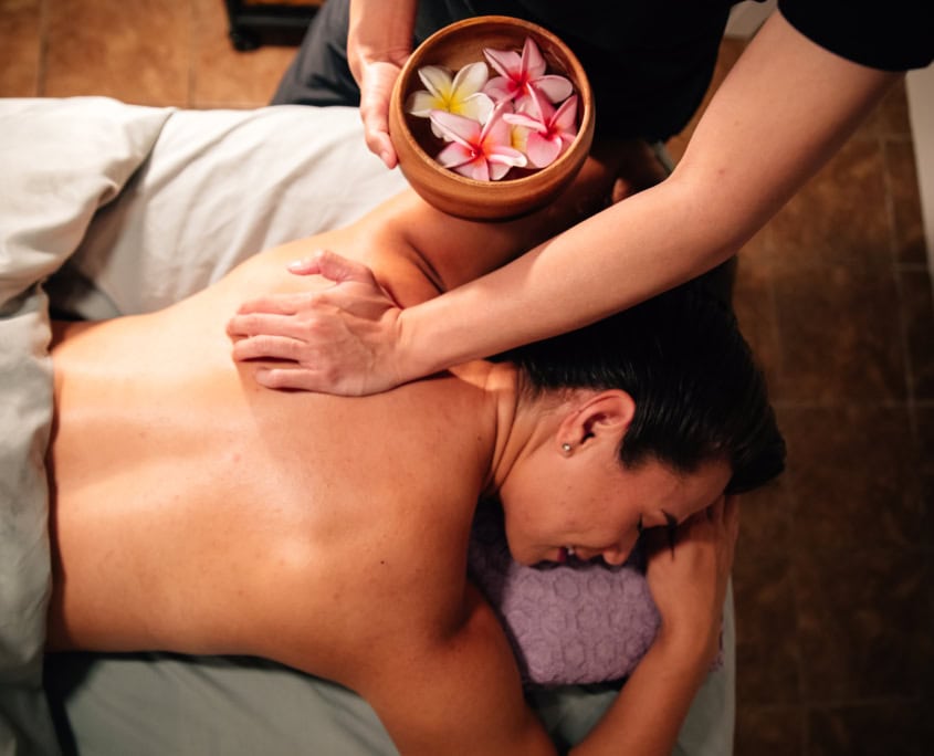 A woman, face down, getting a massage. The masseuse is holding a bowl of plumeria flowers.