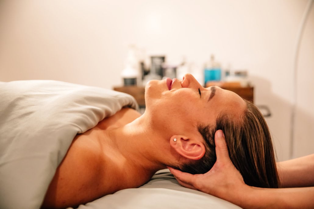 A woman with her eyes closed wearing a white towel receiving a cranial massage.