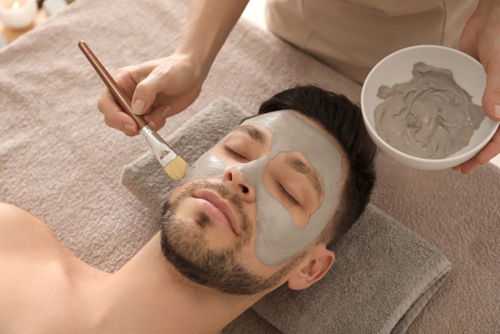 A man with a beard receiving a skin treatment as a green paste is applied to his face with a brush